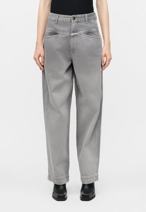 Άτομο που φοράει γκρι τζιν παντελόνι ψηλόμεσο, φαρδύ, με μπροστινές τσέπες και μαύρες μποτάκια αστραγάλου, στέκεται μπροστά σε ένα απλό φόντο.