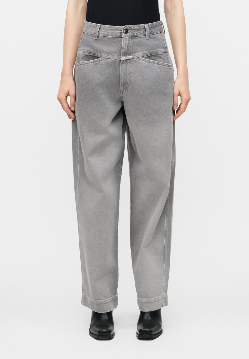 Άτομο που φοράει γκρι τζιν παντελόνι ψηλόμεσο, φαρδύ, με μπροστινές τσέπες και μαύρες μποτάκια αστραγάλου, στέκεται μπροστά σε ένα απλό φόντο.