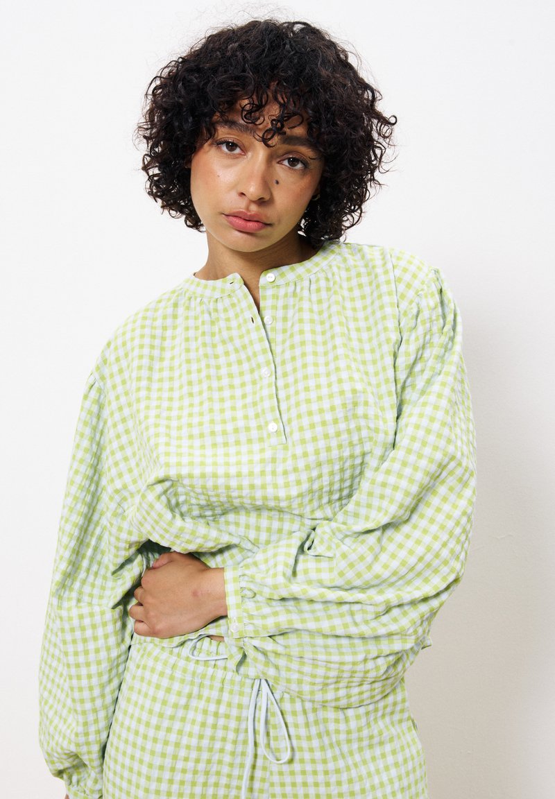 Chemise à carreaux verts avec un col rond et une patte de boutonnage, manches bouffantes longues, poignets froncés et ourlet à cordon. Tissu doux et texturé.