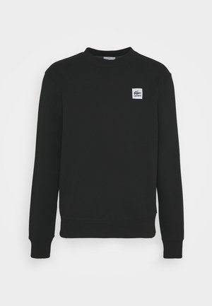 Person, die ein schwarzes Langarmshirt mit kleinem weißem Logo, dunkelblaue Jeans und weiße Sneakers trägt, vor einem schlichten hellen Hintergrund stehend.