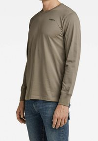 Mężczyzna w dopasowanej, taupe'owej koszuli z długim rękawem z małym czarnym logo na klatce piersiowej, w połączeniu z niebieskimi dżinsami, na białym tle.