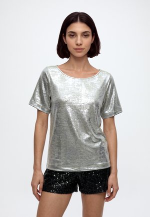 Femme aux cheveux courts et foncés portant un haut argenté brillant à manches courtes et un short noir pailleté sur un fond blanc uni.