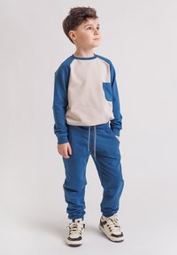 Chłopiec stojący i patrzący w górę w prawo, ubrany w beżowo-niebieską koszulę z długim rękawem, niebieskie spodnie dresowe i białe tenisówki z czarnymi akcentami.