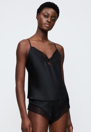 Junge Frau trägt schwarzen Satin-Camisole und passende Shorts mit Spitzenbesatz, stehend vor schlichtem hellgrauem Hintergrund.