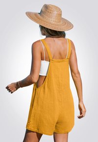 Achteraanzicht van een vrouw die een breedgerande strohoed, een wit crop top en een mosterdgele mouwloze romper met zijknips draagt.
