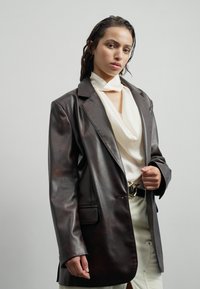 Sacou din piele maro cu rever în vârf, buzunare frontale și o textură netedă, purtat peste un top crem drapat și pantaloni deschiși.