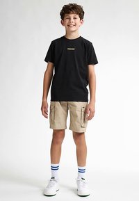 Musta lyhythihainen t-paita, jossa kultainen logo, yhdistetty beigen värisiin cargo-shortseihin. Käyttäjällä on valkoiset tennarit, joissa vihreitä yksityiskohtia, ja siniraidalliset sukat.