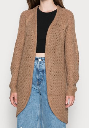 Femme portant un long cardigan marron en maille ouvert sur le devant, un crop top noir et un jean bleu taille haute avec fermeture à boutons.