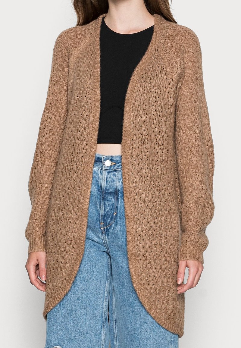 Femme portant un long cardigan marron en maille ouvert sur le devant, un crop top noir et un jean bleu taille haute avec fermeture à boutons.