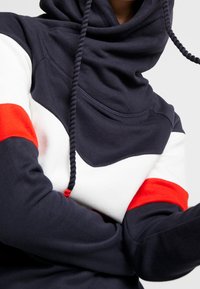 Persona che indossa un hoodie blu scuro con maniche colorate in bianco e rosso e corde spesse blu scuro, su uno sfondo semplice.