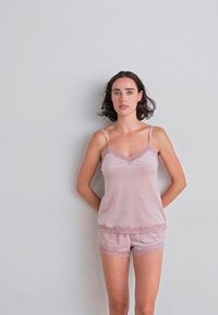 Modelka v růžovém krajkovém topu a kraťasech, stojící na světle šedém pozadí, s rukama položenýma za zády.