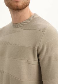 Beige kudumisvillane, millel on tekstuuriga triibud, ümmargune kaelus ja sile pind. Kanga kaudu on näha kudumise variatsioone ja kerget läiget.