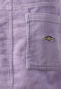 Fialový kordový materiál s vertikální strukturou, obsahující náprsní kapsu a vyšívané logo ve tvaru diamantu na povrchu.