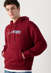 Burgundy hoodie made of cotton blend, featuring front pocket, drawstring hood, and white logo text "Calvin Klein NEW YORK" across chest.