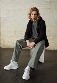 Svart fleecejacka, grå t-shirt, ljusgröna cargobyxor, vita sneakers. Modell sitter på en geometrisk block mot en strukturerad vägg.