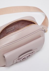 Borsa a spalla in pelle rosa chiara con texture liscia, zip dorata e un elemento decorativo in metallo sulla parte anteriore. Dotata di tracolla regolabile.