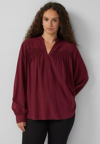 Bordeauxkleurige blouse met lange mouwen en een diepe halslijn, versierd met smockdetails op de schouders, gemaakt van een gladde stof. Gemaakt in combinatie met zwarte broeken.
