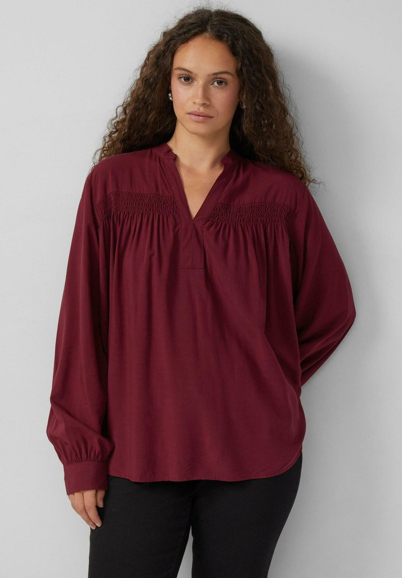 Bordeauxkleurige blouse met lange mouwen en een diepe halslijn, versierd met smockdetails op de schouders, gemaakt van een gladde stof. Gemaakt in combinatie met zwarte broeken.