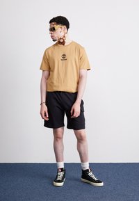 Ērta īsām piedurknēm t-kreklā ar melnu Timberland logo, apvienots ar melnām šortiem un augstiem melniem sporta apaviem ar krēmkrāsas zolemi un baltām zeķēm.