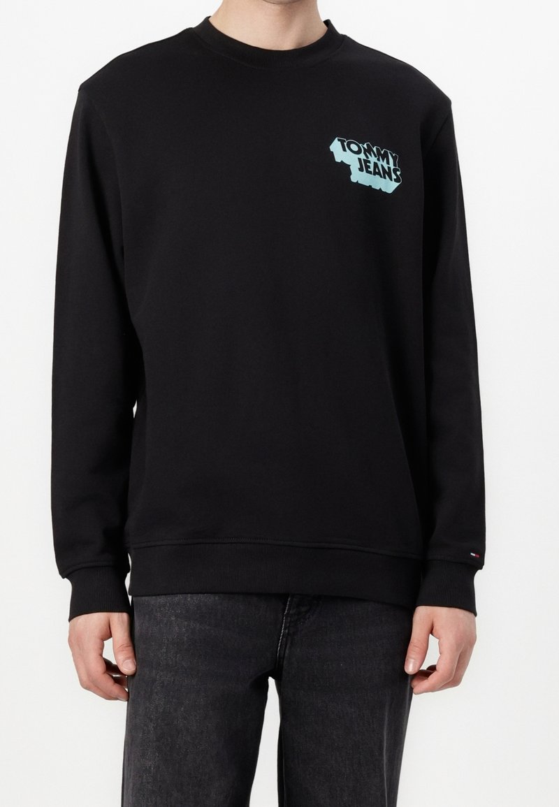 Personne portant un sweat-shirt noir à manches longues avec le logo "Tommy Jeans" bleu clair sur la poitrine gauche et un jean gris foncé.