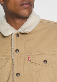 Giacca beige in velluto a coste con colletto in pelliccia sintetica color crema, caratterizzata da bottoni metallici e una piccola etichetta rossa Levi's sulla tasca frontale.