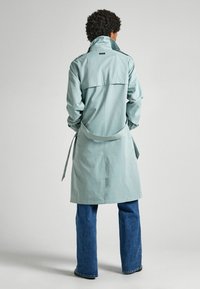 Jasny płowy trencz z taliowaniem, wysokim kołnierzem i długimi rękawami, noszony na niebieskich rozszerzanych dżinsach. Gładka faktura, z detalem w postaci paska.