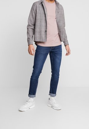 Hombre con chaqueta de cuadros gris, camiseta rosa, vaqueros remangados azules, calcetines blancos y zapatillas blancas de pie contra un fondo gris liso.