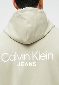 Ανοιχτό μπεζ φούτερ με μεγάλο λευκό κείμενο "Calvin Klein JEANS" στο πίσω μέρος. Διαθέτει κουκούλα με κορδόνι και μαλακό, υφασμάτινο υλικό.