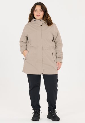 Kvinde iført beige hættejakke med lommer, sorte bukser og sorte sneakers, stående mod en ensartet lys baggrund.