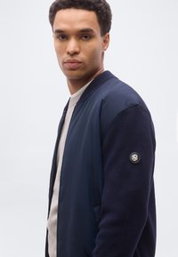 Νέος άνδρας που φοράει ναυτικό μπουφάν τύπου bomber πάνω από ανοιχτό μπεζ πουλόβερ, στέκεται μπροστά σε ένα απλό φωτεινό φόντο, κοιτάζοντας ελαφρώς προς το πλάι.