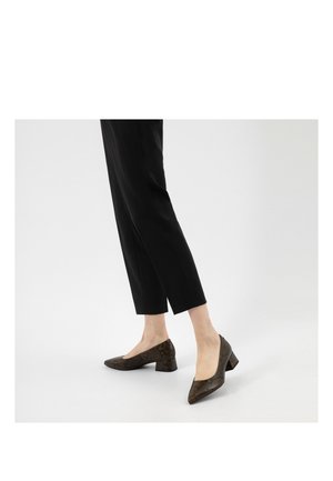 Vrouw draagt zwarte cropped broek en bruine lage blokhak schoenen met slangenhuidprint, staand op een witte vloer.