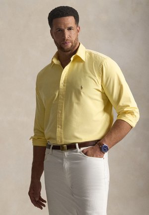 Homme portant une chemise jaune à boutons avec manches retroussées, pantalon blanc, ceinture marron et montre-bracelet bleue, debout avec une main dans la poche.