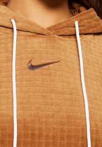 Brun vadderad hoodie med vita snörningar; har en liten brun Nike-logga på bröstet; mjukt, texturerat tyg.