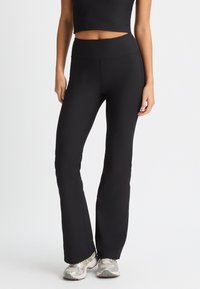 Leggings negros acanalados de corte acampanado con una cintura alta, textura suave y silueta ajustada, combinados con zapatillas deportivas plateadas.