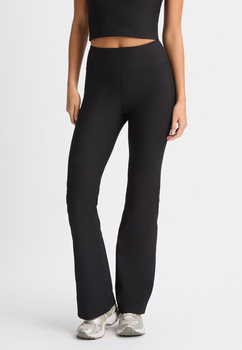 Leggings negros acanalados de corte acampanado con una cintura alta, textura suave y silueta ajustada, combinados con zapatillas deportivas plateadas.