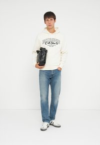 Beige hættetrøje med trykt logo, blå denim jeans, sort taske med spænder og hvide sneakers med sorte accenter. Ensfarvet baggrund.