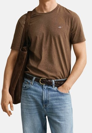Homme portant un t-shirt marron à manches courtes, un jean bleu clair avec une ceinture tressée marron foncé, et un sac à épaule marron assorti.