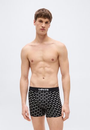 Άνδρας χωρίς πουκάμισο, φοράει μαύρα μπόξερ Levi's με λευκό λογότυπο, στέκεται μπροστά σε ένα απλό ανοιχτό φόντο.