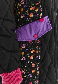 Prešívaná čierna bunda s kvetinovou vnútornou podšívkou. Má fialový chlopňu so svetloružovým cvokom a textúrované ružové pletené manžety.