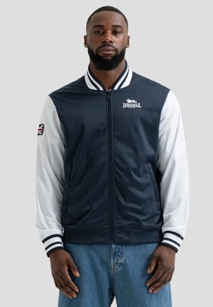 Homme barbu portant une veste zippée Lonsdale London bleu marine et blanc avec un patch Union Jack sur la manche et un jean bleu, debout devant un fond gris.