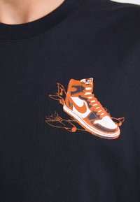 T-shirt noir avec un graphisme orange d'une sneaker montante, avec des accents et des lacets blancs, imprimé sur la zone de la poitrine.