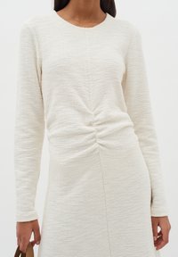 Vit långärmad klänning med en strukturerad tyg. Har en vriden frontknytning och en rak silhuett.