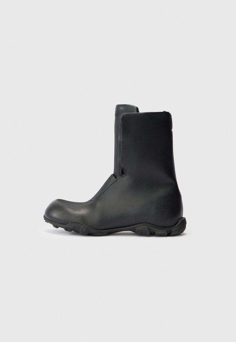 Bottine noire en matériau lisse semblable au caoutchouc, dotée d'un bout arrondi, d'un design minimaliste et d'une semelle texturée avec des motifs de traction.