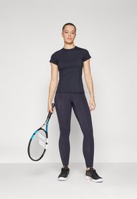 T-shirt atlético preto de mangas curtas combinado com leggings pretas de cintura alta, segurando uma raquete de ténis, sapatilhas atléticas pretas com detalhes texturizados.
