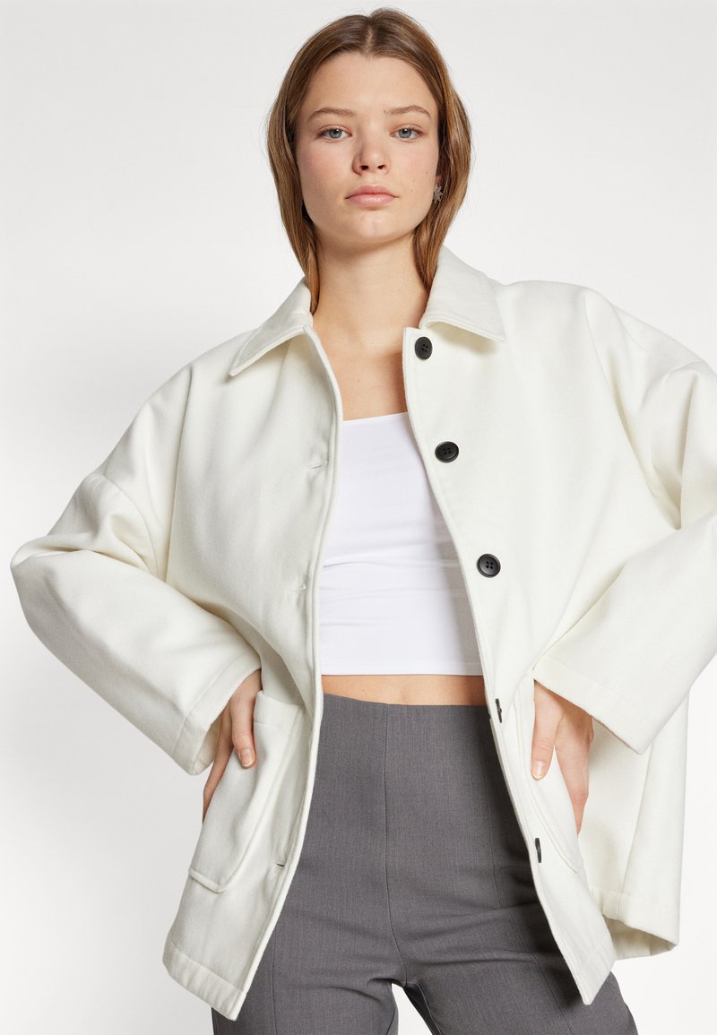 Chaqueta blanca de gran tamaño hecha de tejido suave, con cuello, botones frontales y bolsillos laterales. Llevada sobre una blusa blanca y pantalones grises.