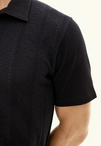 Polo shirt lavorato a maglia nera con un motivo a diamante testurizzato sulla parte anteriore, maniche corte e colletto classico. Finitura in tessuto morbido.