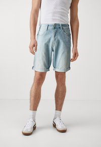 Pantalones cortos de mezclilla azul claro con dobladillos enrollados, diseño de cinco bolsillos y cierre de botón. Llevados con zapatillas blancas y calcetas hasta los tobillos.