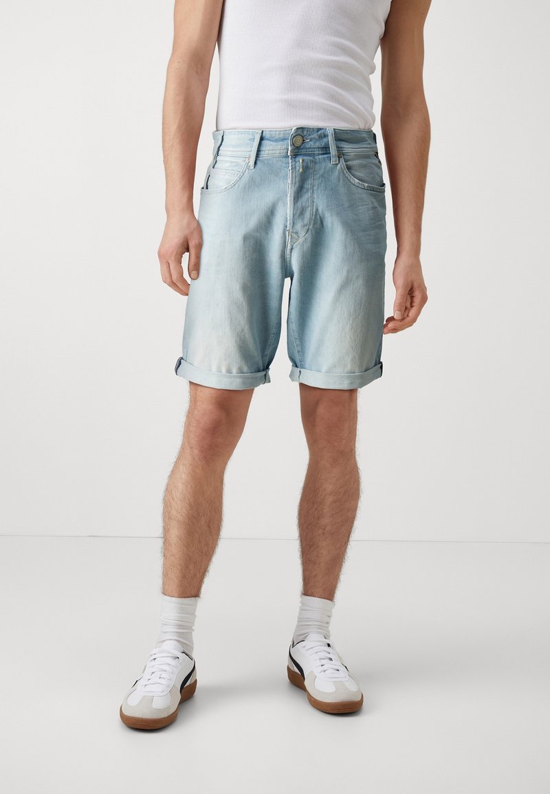 Pantalones cortos de mezclilla azul claro con dobladillos enrollados, diseño de cinco bolsillos y cierre de botón. Llevados con zapatillas blancas y calcetas hasta los tobillos.