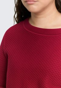Bordeauxfarbener Pullover aus strukturiertem, gesteppter Stoff mit einem gerippten Rundhalsausschnitt. Nahaufnahme, die das Stoffmuster und die Nähdetails zeigt.