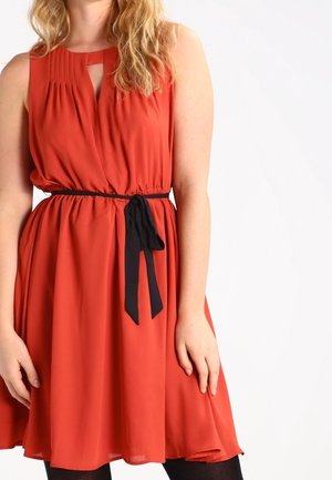 Femme portant une robe sans manches orange rouille avec des plis, un décolleté en forme de trou de serrure et une ceinture noire nouée à la taille, sur un fond uni.